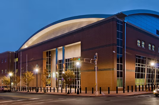 A modern brick and glass building with an arched roof and bulletproof windows is illuminated at dusk.