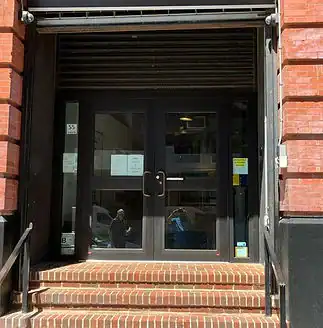 Glass double bullet resistant doors with signs set in brick at the top of a short railed staircase.