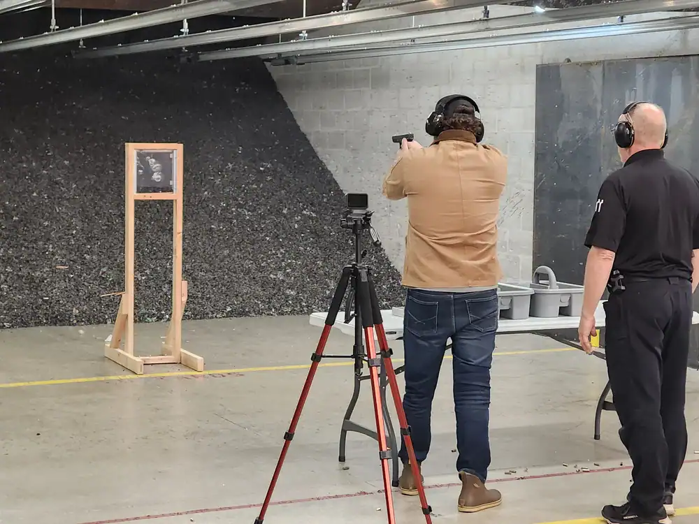 Two people at an indoor shooting range, one aiming a handgun at a target as ballistic doors stand behind.