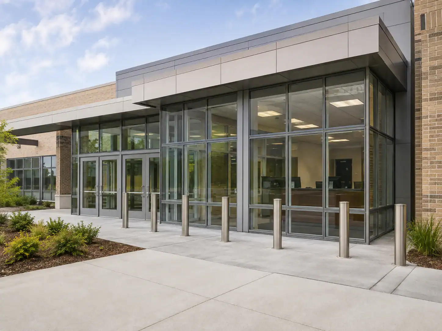 A modern building entrance features ballistic doors, glass windows, bollards, and landscaping outside.